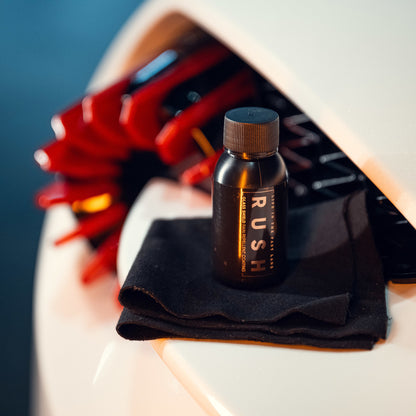 Bovenop een witte auto met rode accenten staat een klein zwart flesje met de tekst RUSH op een gevouwen zwarte doek - perfect voor het aanbrengen van autoglascoating.