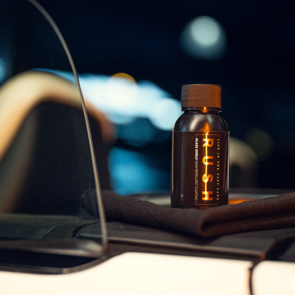 Een klein flesje met het label RUSH ligt op een opgevouwen zwarte doek in een auto, naast een raam dat is behandeld met waterafstotende ruitencoating.