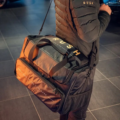A person in a black jacket carries a large black Detailing Bag with RUSH branding and "LIVE IN THE FAST LANE" on the side—ideal for your Detailing Package PLUS | 23-delig or any auto reinigingspakket essentials. Features shoulder and hand straps.
