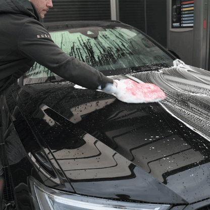 Persoon die een zwarte auto wast met autoshampoo en een zeepspons, die schuim verspreidt voor extra winterbescherming auto bij een wasstraat.
