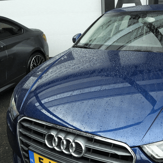 Een blauwe Audi met regendruppels op de motorkap staat geparkeerd bij een grijze auto - perfect voor het testen van een microvezel droogdoek of droogdoek 600 GSM.