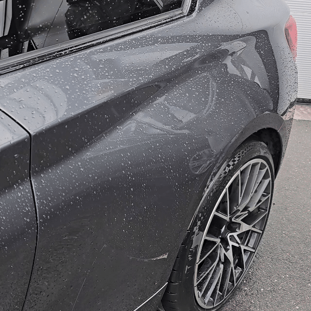 Close-up of a black car’s rear side and wheel, covered in raindrops. Treated with the Black Friday Package, the car gleams on a paved surface as reflections play across its glossy finish.