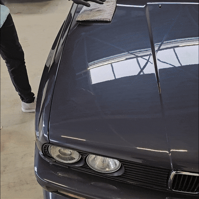 A person wearing a glove is polishing the hood of a dark-colored car with 1+1 GRATIS Spray Wax for lak bescherming in a garage. Only their arm and part of one leg are visible. The car's headlights and reflections on the hood stand out.