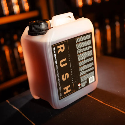 Een plastic 2,5-liter vat met de tekst RUSH Iron Remover for Wheels and Bodywork staat op een donker oppervlak in een schemerige kamer.