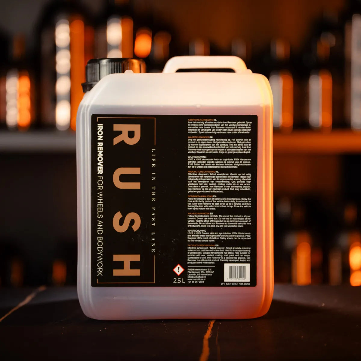 Een 2,5-liter vat met de naam Rush Iron Remover for Wheels and Bodywork op een donker oppervlak met vage flessen op de achtergrond.