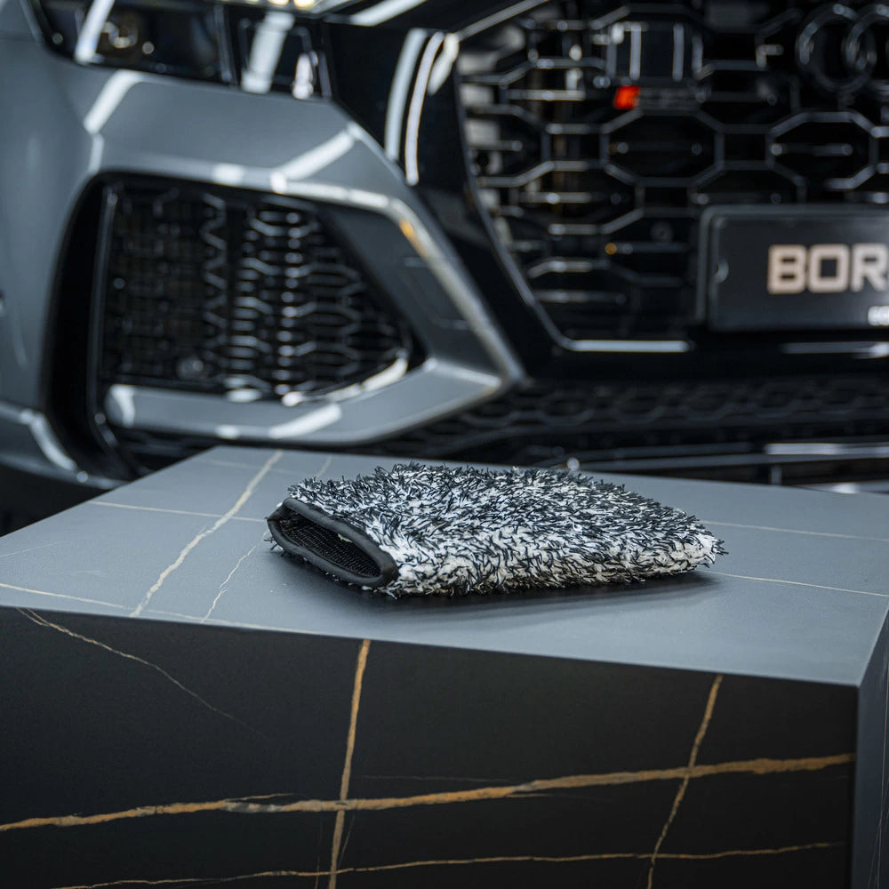 Een microvezel washandschoen met groot absorberend vermogen rust op een grijze doos voor de zichtbare grille en koplamp van een geparkeerde auto.