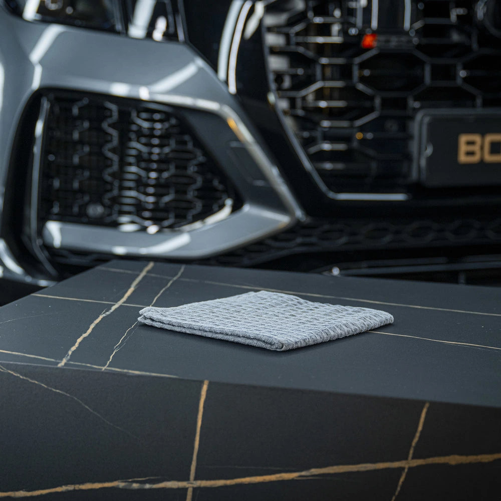 Een grijs microvezeldoek ligt op een zwarte tafel met gouden adering, met de grille van een grijze auto op de achtergrond.