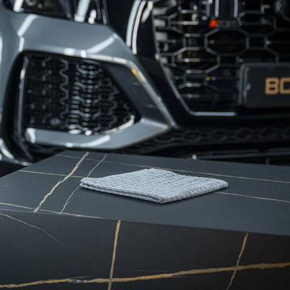 Een grijs microvezeldoek ligt op een zwarte tafel met gouden adering, met de grille van een grijze auto op de achtergrond.