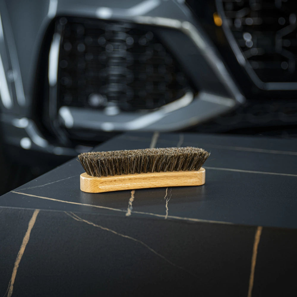 The Leather Cleaning Brush with dark bristles rests on a black, marble-like surface with gold veins, while a blurred car front grille and headlight appear in the background.