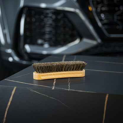 The Leather Cleaning Brush with dark bristles rests on a black, marble-like surface with gold veins, while a blurred car front grille and headlight appear in the background.