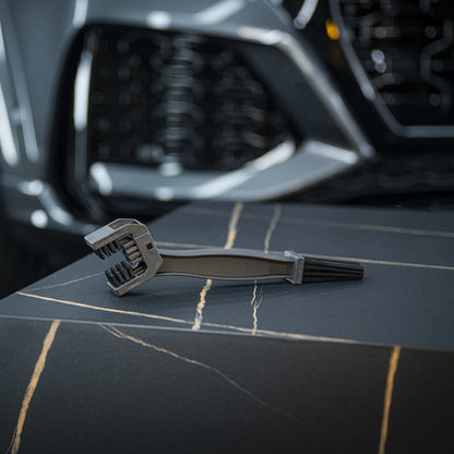 A Chain Brush made of black plastic sits on a dark, marble-patterned surface, while a car's front grille and headlight appear blurred in the background.