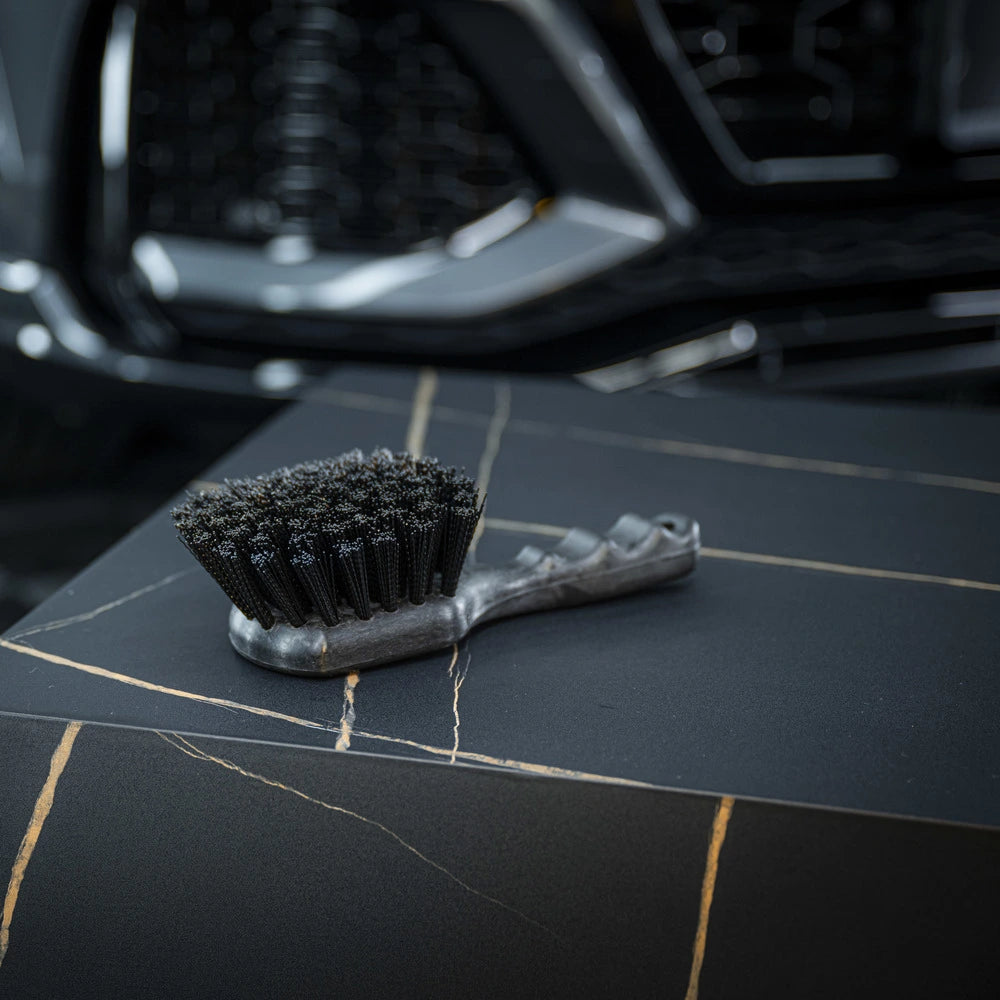 Een kleine harde borstel met stugge haren rust op een donker, gemarmerd oppervlak; op de achtergrond is een autogrille zichtbaar.