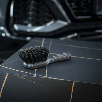 Een kleine harde borstel met stugge haren rust op een donker, gemarmerd oppervlak; op de achtergrond is een autogrille zichtbaar.