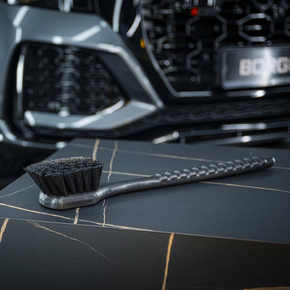 Een harde borstel rust op een donker oppervlak met marmerpatroon; op de achtergrond is een grille van een auto zichtbaar.