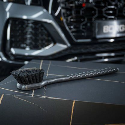 Een harde borstel rust op een donker oppervlak met marmerpatroon; op de achtergrond is een grille van een auto zichtbaar.