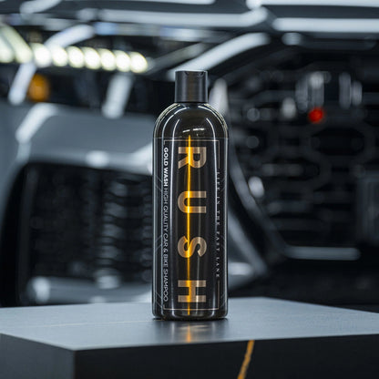 Een zwarte PUSH-fles voor autoshampoo staat op een donker oppervlak, met een wazige voorkant van een auto op de achtergrond, klaar om te eikelen en te autowassen.
