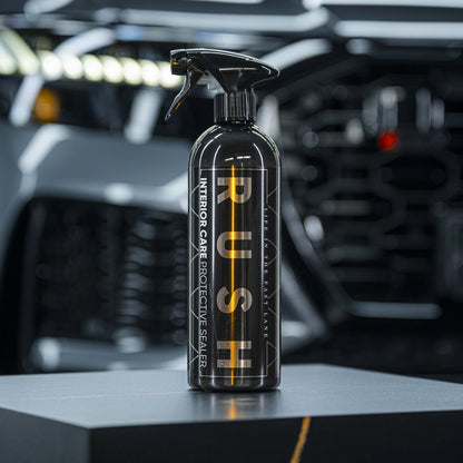 Een zwarte interieurverzorgingsspray met het label PUSH staat op een donker oppervlak, met daarachter een wazig auto-interieur, ideaal voor interieur beschermen auto.