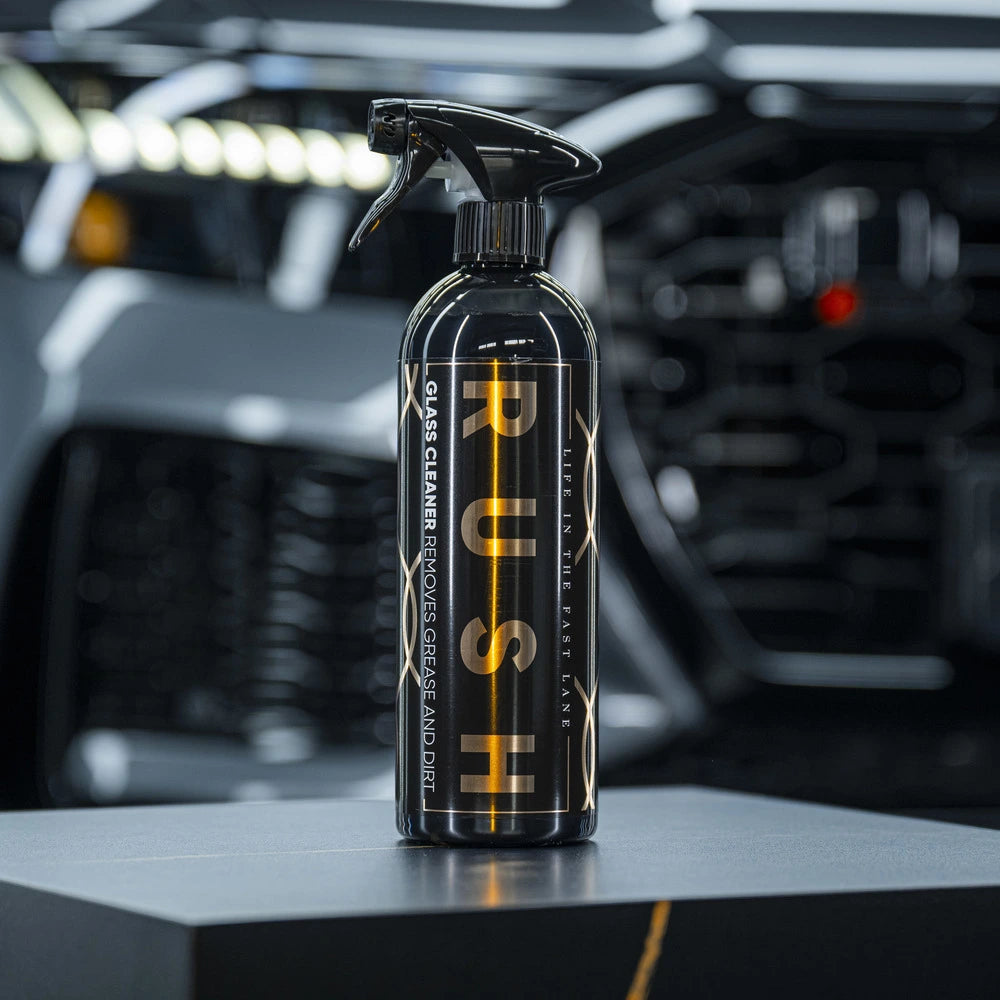 Een zwarte RUSH Glasreiniger spuitfles staat op een donker oppervlak met een wazig voertuig op de achtergrond, perfect voor streepvrij glas reinigen.