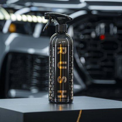 A black spray bottle labeled Ceramic Sealant, noted for its keramische lakbescherming, stands on a dark surface with a blurred car grille and headlights in the background, accentuating the bottle’s glossy finish and sleek modern design.