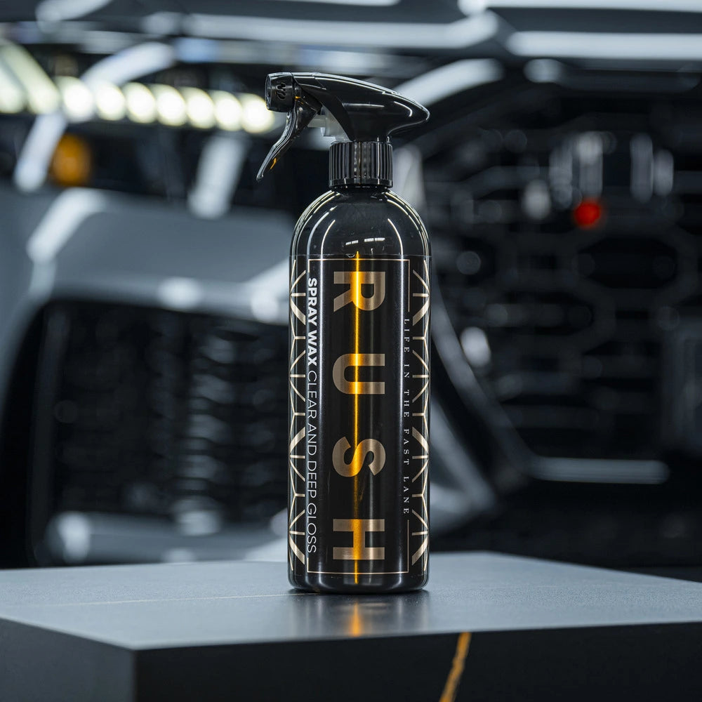 A bottle of 1+1 GRATIS Spray Wax sits on a dark surface, labeled PUSH SPRAY WAX CLEAR AND DEEP GLOSS, with lak bescherming and advanced nanotechnologie, against a blurred car and automotive-themed background.