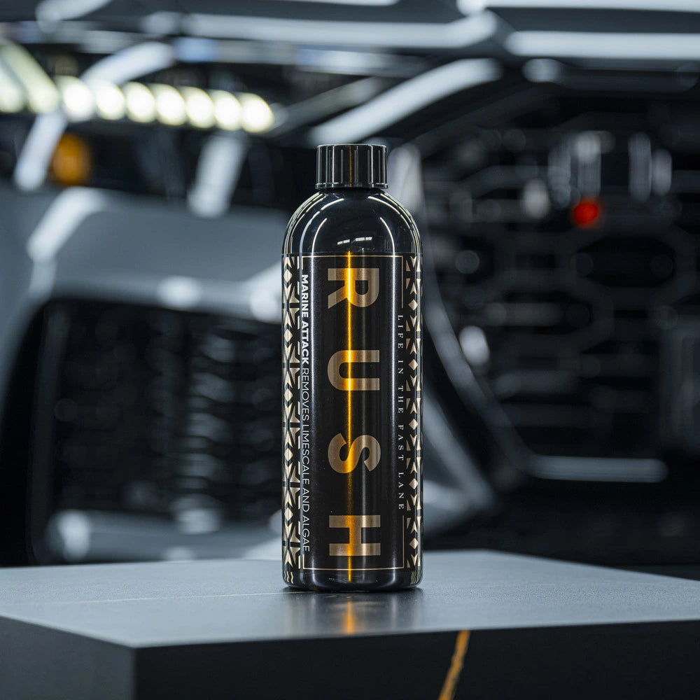 A sleek black bottle labeled 1+1 GRATIS Marine Attack stands on a dark surface, with a blurred modern car behind. Perfect for boot reinigen, it's made from biologisch afbreekbaar material and features a geometric side pattern.