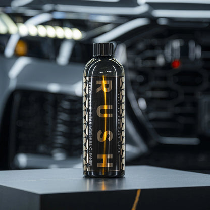 Een zwarte fles met het label PUSH Extreme Wheel  Multi Cleaner voor bootreiniging staat op een grijs oppervlak met een vage auto op de achtergrond.
