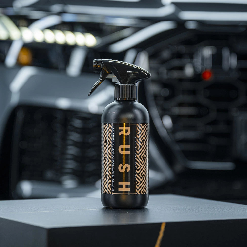 A black spray bottle labeled Leather Matt Clean with a geometric gold and black pattern stands on a dark surface, perfect for lederen autobekleding reinigen, with a blurred modern car grille and headlights in the background.