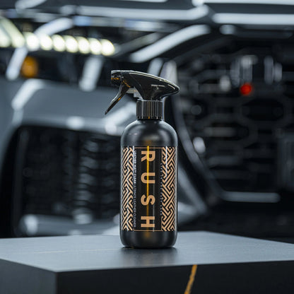 A black spray bottle labeled Leather Matt Clean with a geometric gold and black pattern stands on a dark surface, perfect for lederen autobekleding reinigen, with a blurred modern car grille and headlights in the background.