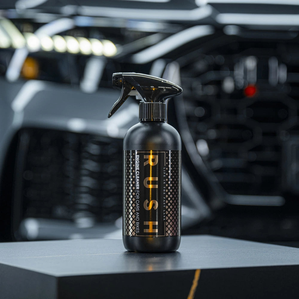 Een zwarte spuitfles met de tekst PUSH, krachtige alkalische reiniger voor motorruimte reiniging, staat voor een vage grille en koplampen.