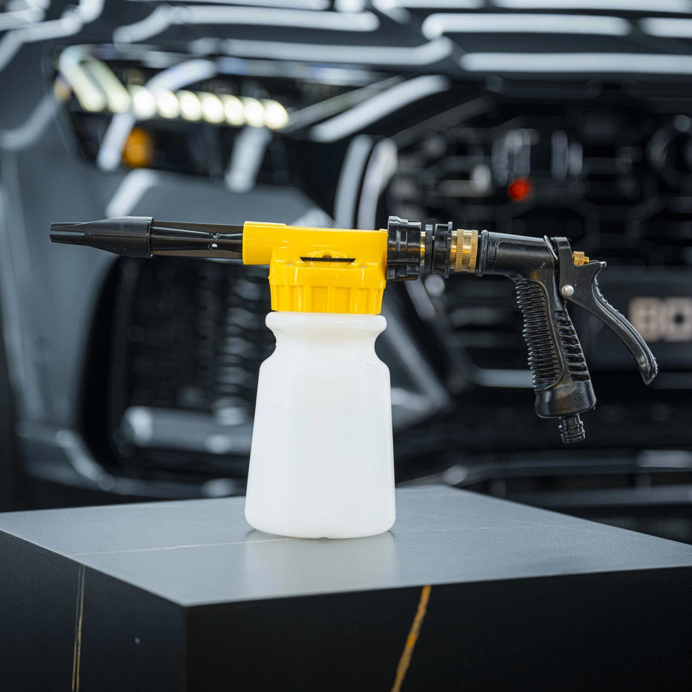 A black and yellow Foam & Water Spray Gun with a white soap bottle is displayed on a dark pedestal, the blurred front grill of a black vehicle in the background—perfect for use without a pressure washer.