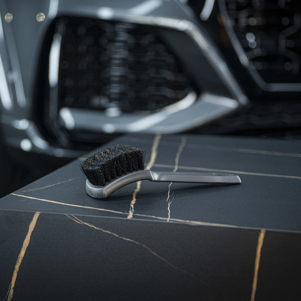 The Tire Brush, small and black with a silver handle, rests on a dark marble-like surface streaked with light tones. A blurred car front is visible in the background.