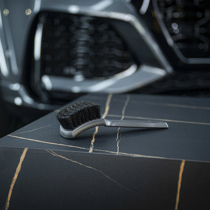 The Tire Brush, small and black with a silver handle, rests on a dark marble-like surface streaked with light tones. A blurred car front is visible in the background.