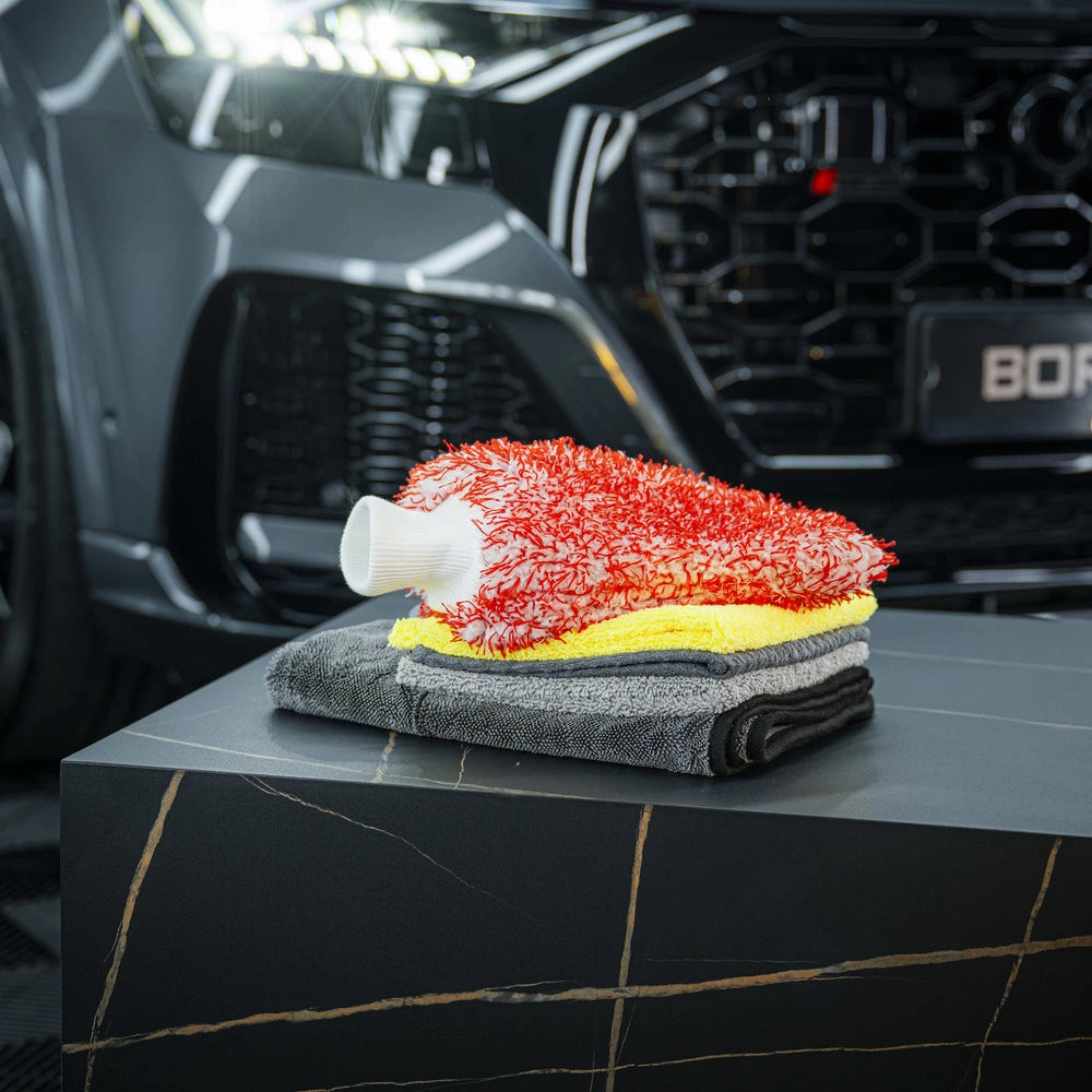 Een rode Microfiber Wash Mitt en twee gevouwen Drying Towel 600 GSM autoaccessoires rusten op een marmeren oppervlak voor een zwarte auto.