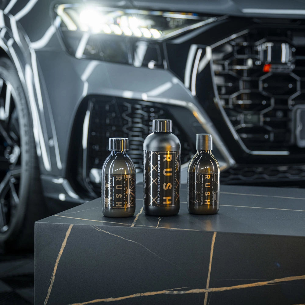 Drie zwarte flessen - RUSH Spray Wax, RUSH Bug Remover en RUSH Gold Wash - staan op een donker marmeren oppervlak voor een gestroomlijnde zwarte auto.