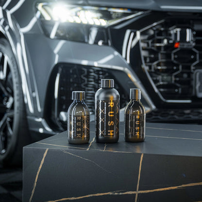 Drie zwarte flessen - RUSH Spray Wax, RUSH Bug Remover en RUSH Gold Wash - staan op een donker marmeren oppervlak voor een gestroomlijnde zwarte auto.