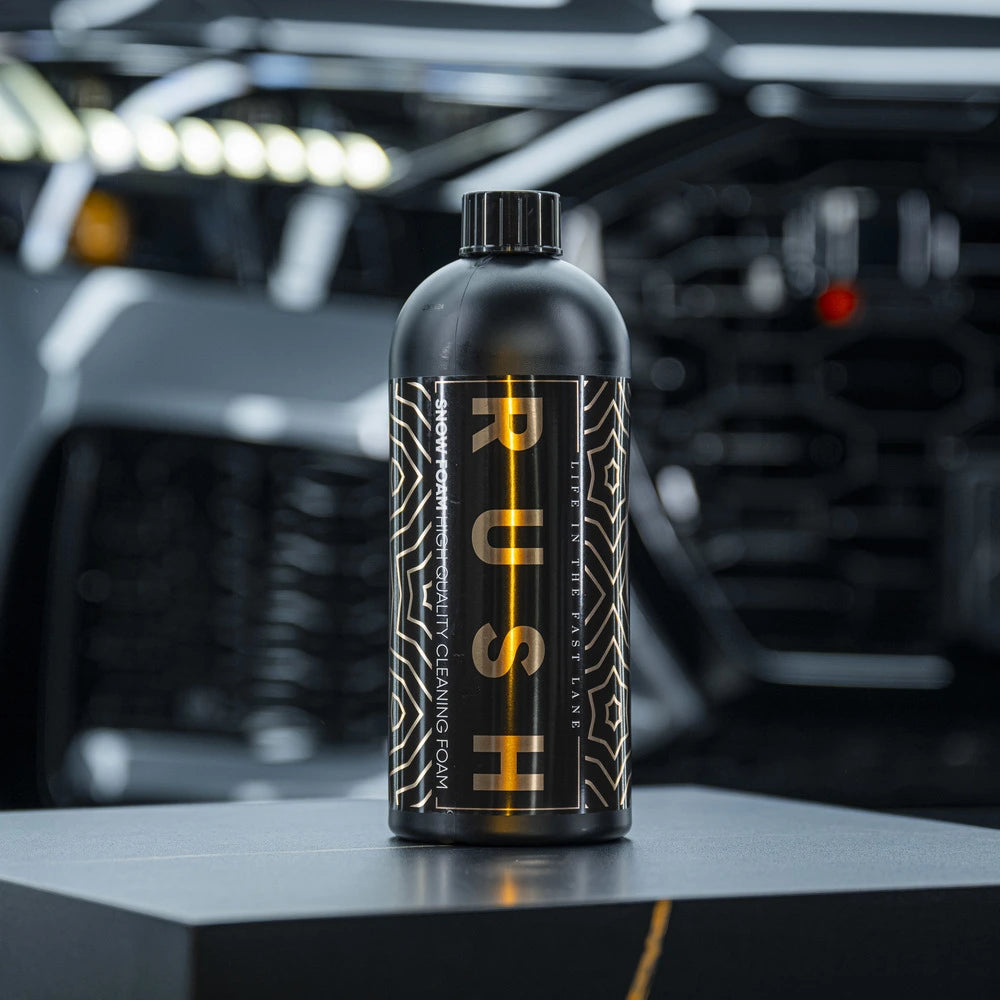 Een zwarte RUSH-fles voor autovoorwas staat op een grijs oppervlak, met een wazige auto en felle lichten op de achtergrond.