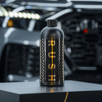 Een zwarte RUSH-fles voor autovoorwas staat op een grijs oppervlak, met een wazige auto en felle lichten op de achtergrond.