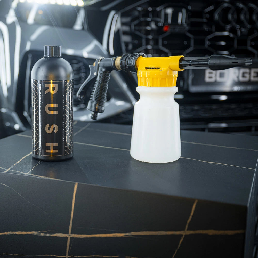 A black RUSH bottle and a yellow-and-white Foam Spray Gun from the Foam Spray Gun Package rest on a dark surface before a modern car grille, ready to deliver the perfect snow foam auto voorwas.
