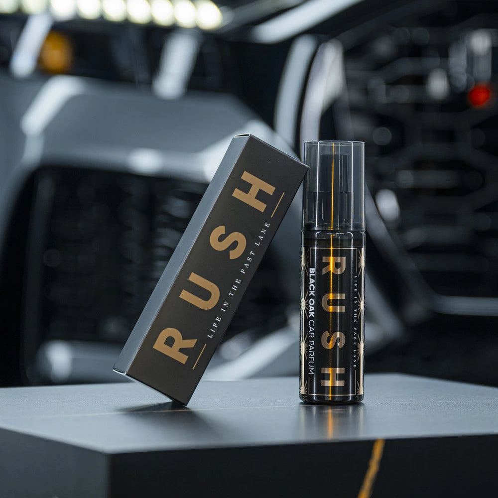 Een zwarte autoparfumfles met het label RUSH en het doosje, beide met gouden tekst, voor een vage grille van een auto.