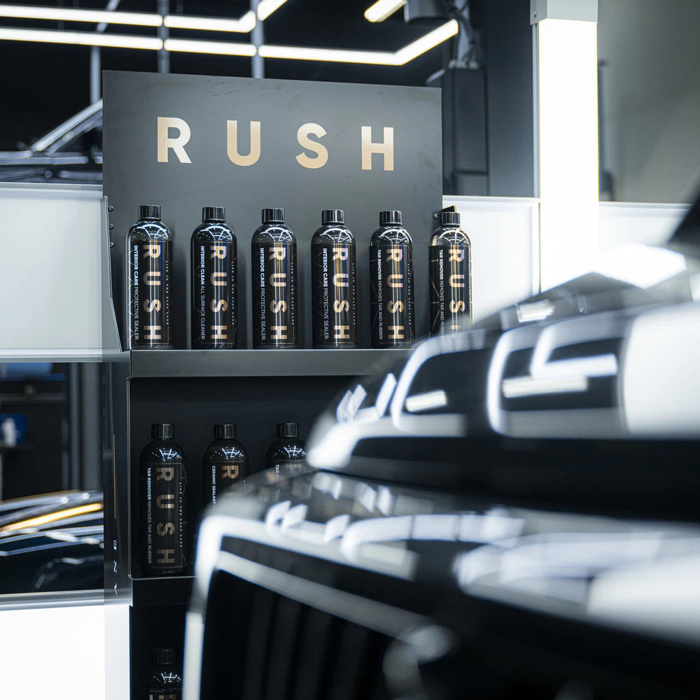 Neatly arranged on shelves in a modern, well-lit space are black bottles labeled RUSH—part of the Starter Kit | 21-delig. The word RUSH glows above, while the front of a glossy black vehicle appears in the foreground.