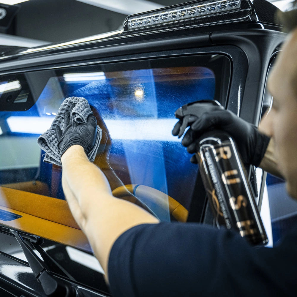 Een persoon met handschoenen aan maakt een autoruit schoon met glasreiniger en een microvezeldoek voor een streeploze afwerking.