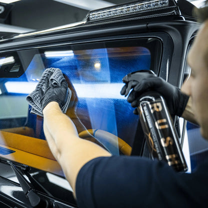 Een persoon met handschoenen aan maakt een autoruit schoon met glasreiniger en een microvezeldoek voor een streeploze afwerking.