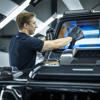 A man wearing black gloves cleans the rear window of a black vehicle using a cloth and cleaning spray from the Starter Kit XXL | 25-delig in a well-lit garage.