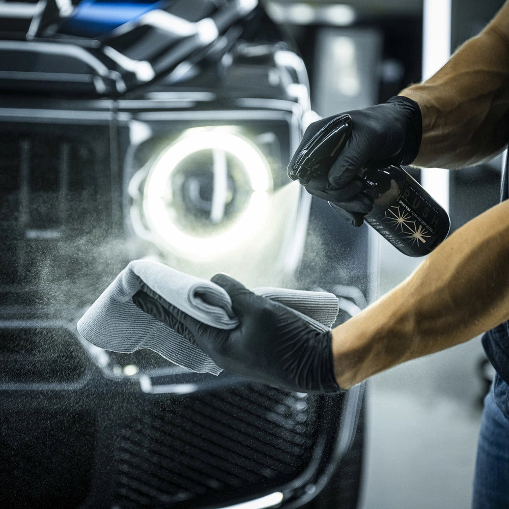 A person wearing black gloves sprays cleaner from the Starter Kit | 21-delig onto a gray microfiber cloth in front of a black car with a round headlight, preparing to autopoetsen the vehicle.