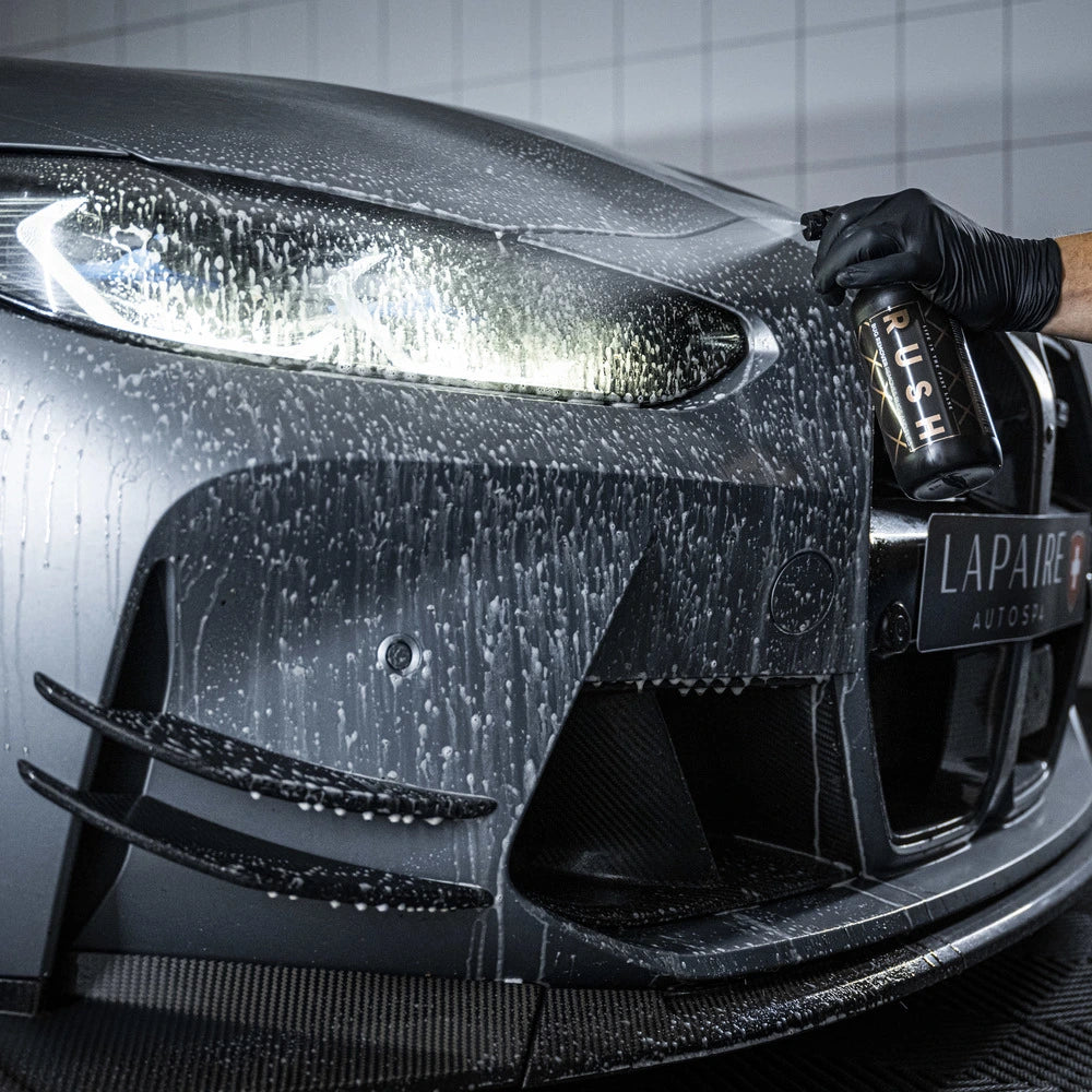 A close-up of a wet, gray sports car’s front bumper and headlight shows a gloved hand holding the Starter Kit XXL | 25-delig bottle during expert detailing at Lapare Autospa.