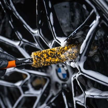 Een gehandschoende hand gebruikt een Wheel Woolie Medium velgenborstel om een zeepachtige, zwarte BMW velg met gele en zwarte details schoon te maken.