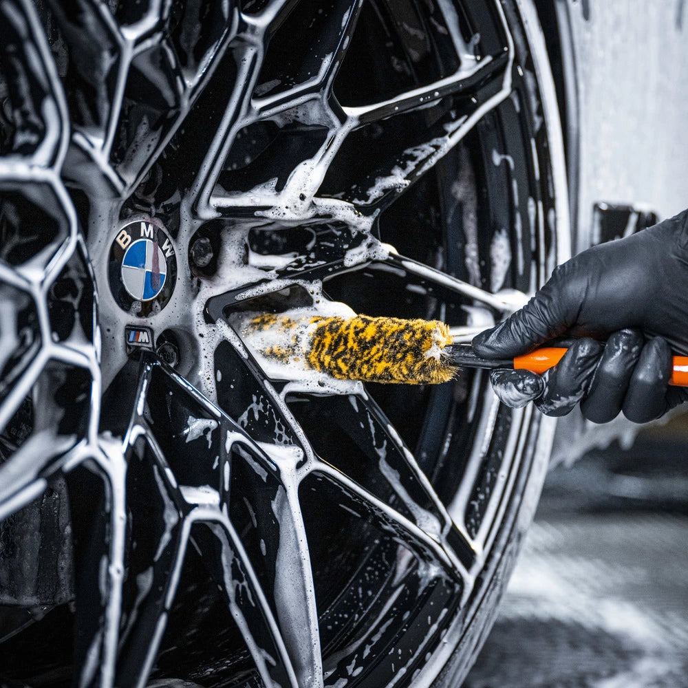 Wearing a black glove, someone uses the Wheel Woolie Small to clean a black BMW wheel, focusing on spaakvelgen reinigen by working soap suds into the intricate spokes and around the BMW logo.