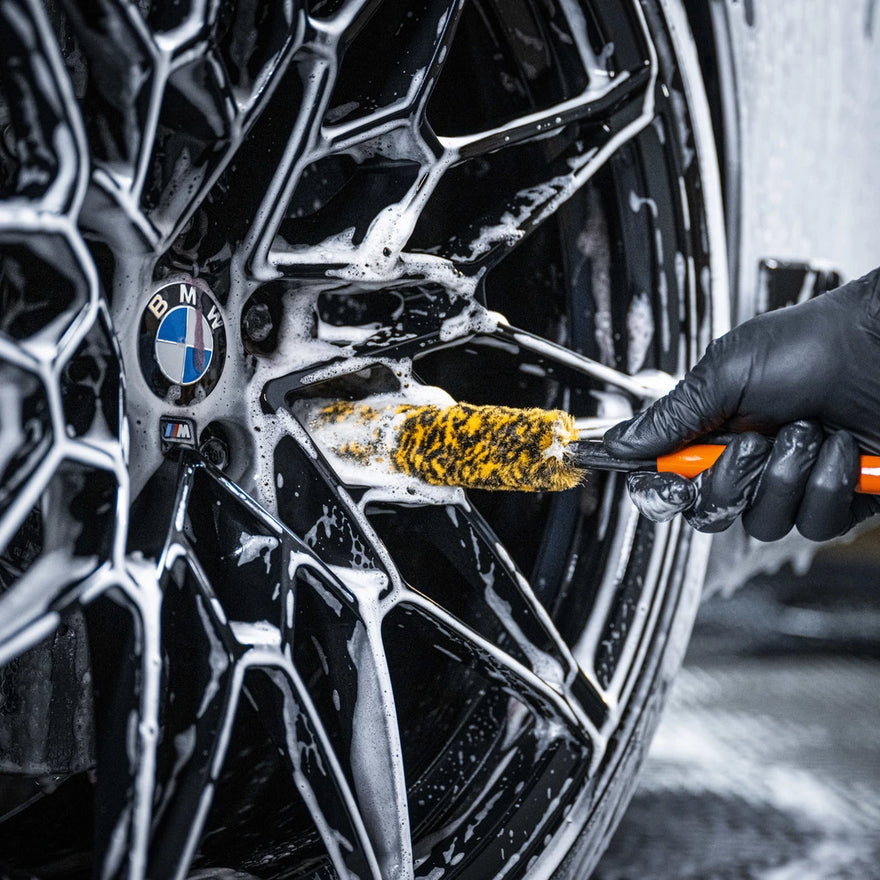 Wearing a black glove, someone uses the Wheel Woolie Small to clean a black BMW wheel, focusing on spaakvelgen reinigen by working soap suds into the intricate spokes and around the BMW logo.