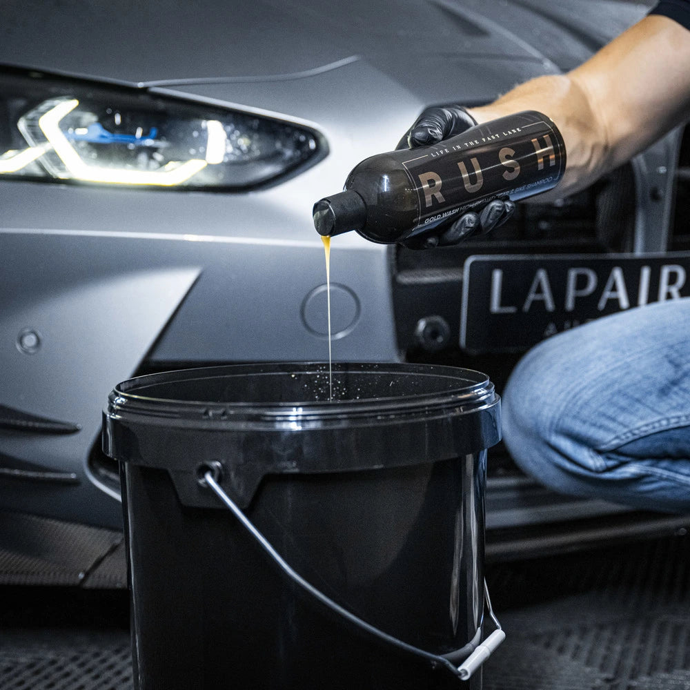 Een persoon giet autoshampoo uit een fles met het label RUSH in een zwarte emmer bij een zilveren auto met de koplampen aan voor extra eikel.