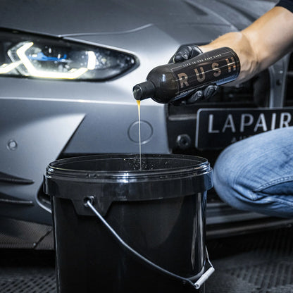 Een persoon giet autoshampoo uit een fles met het label RUSH in een zwarte emmer bij een zilveren auto met de koplampen aan voor extra eikel.